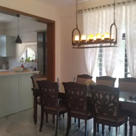 Eight-seater carved dark-wood dining set under a wrought-iron linear candle chandelier, with a sage-green breakfast-counter kitchen open behind it.