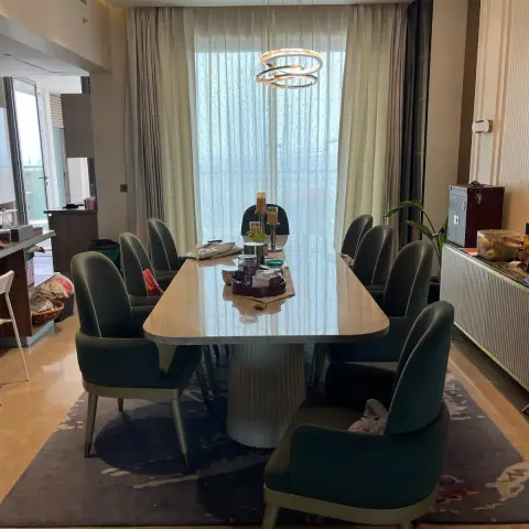 Eight-seater cream marble dining table on a fluted white drum pedestal, ringed by bottle-green leather chairs with brass-tipped legs