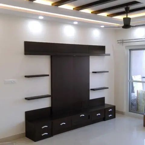 Empty flat with a wenge TV unit — full-height back panel between staggered floating shelves under a wide overhead box — set below a false ceiling banded