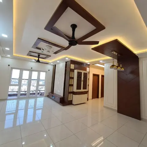 Empty hall with walnut frames set into a cove-lit ceiling over two fans, a carved cream-and-teak pooja cabinet on the far wall