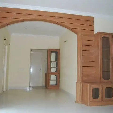 Empty living area with a wood-clad archway on fluted pillars leading to a built-in wood-and-glass crockery display unit along the side wall.