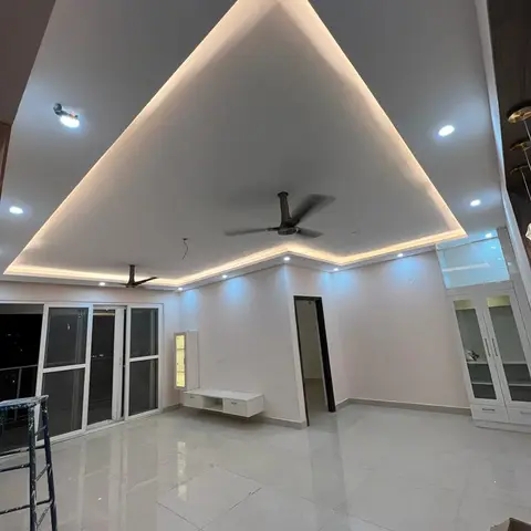 Empty living room mid-fit-out with a dramatic triangular cove-lit ceiling peak, a floating TV console, and a ladder and packing box on the floor.