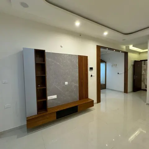 Empty living room with a floating wood-and-stone TV feature wall with open display shelving under a cove-lit false ceiling, opening to a foyer beyond.