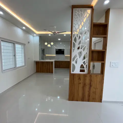 Empty living space with a wood-framed tree-motif jaali partition dividing it from a white modular kitchen beyond, cove-lit ceiling and a ceiling fan overhead