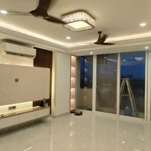 Empty pre-handover hall with a backlit ivory marble TV panel over a floating two-tone console, a square crystal chandelier between two dark wood fans