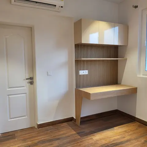 Empty room corner with a floating wood study desk and drawer under a glossy overhead cabinet, backed by a fluted wood-panel niche, wood-look floor tile.