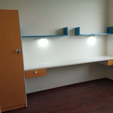 Empty study nook with a floating white desk with orange drawer fronts, blue floating shelves with puck spotlights, and a matching orange wardrobe.