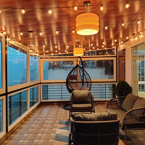 Enclosed balcony sit-out with a hanging egg chair, a rattan sofa set, and warm string lights beneath a wood-plank ceiling.