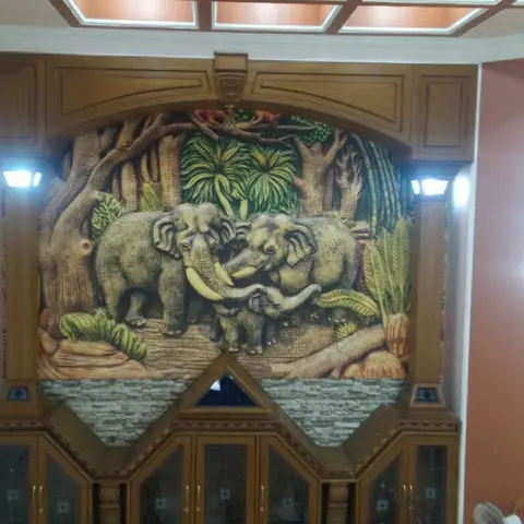 Entrance feature wall with a hand-carved wooden relief mural of elephants set above a glass-fronted display cabinet, flanked by wall sconces.
