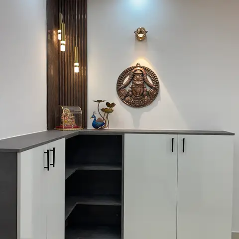 Entrance foyer corner with a fluted wood display niche holding a brass Ganesh idol and peacock figurine, a round brass Balaji wall medallion