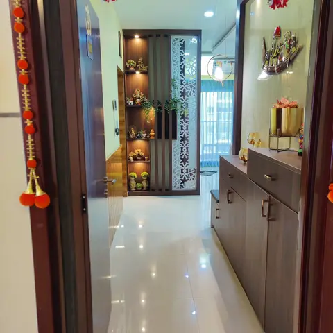 Entrance foyer framed by hanging floral torans, with a backlit wood display shelf of idols and a jaali screen partition on one side