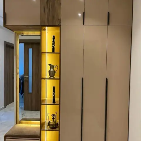 Entrance foyer wall combining a full-height taupe wardrobe with a backlit brass-vase display niche, a mirror, and an upholstered bench with a drawer.