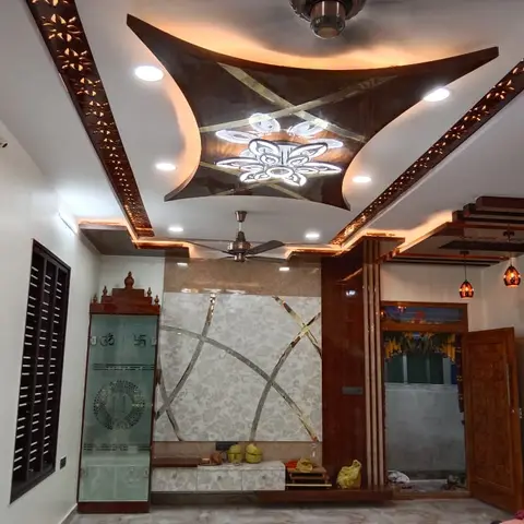 Entrance foyer with a sculpted wing-shaped POP false ceiling with jaali trim, a carved wood main door, and a glass-door pooja cabinet in the corner.