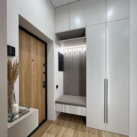 Entrance foyer with a wood-veneer main door, floor-to-ceiling white storage cabinets, and a built-in bench with a fluted panel backdrop and coat hooks.