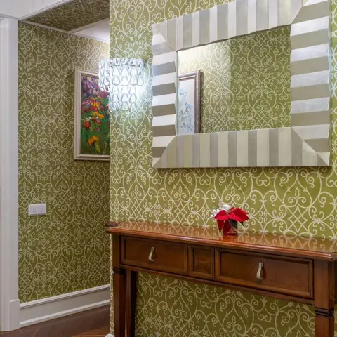 Entrance hallway with ornate olive-green damask wallpaper, a large striped mirror over a wooden console table, and crystal wall sconces.