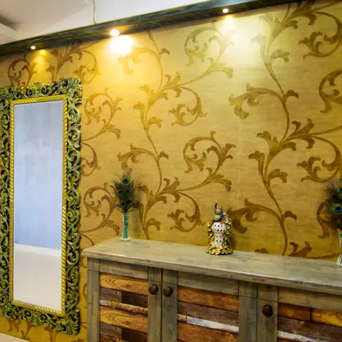 Entrance wall with gold damask wallpaper, an ornate carved mirror and a rustic wood console styled with peacock feathers and a decorative sculpture.