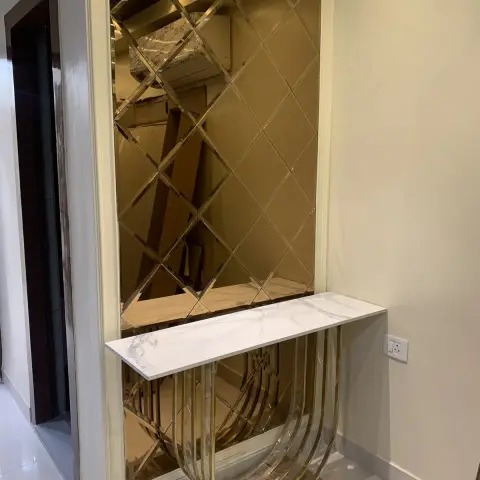Entry console still in its shrink wrap — white marble top on a looping gold metal base against a full-height bronze-tinted bevelled diamond mirror panel