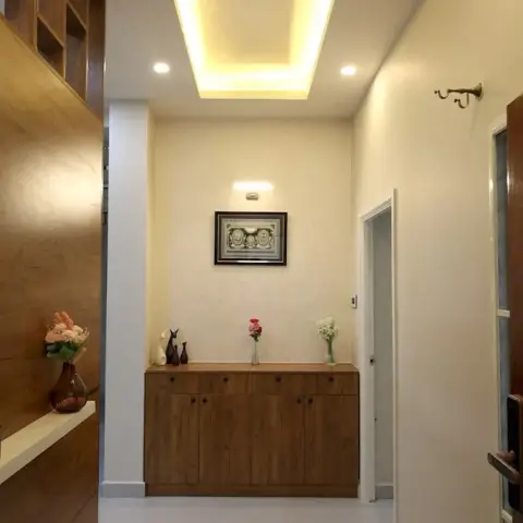Entry foyer with a low wooden shoe cabinet under a framed relief lit by a slim picture light, a wood-clad side wall carrying a white display ledge with plants