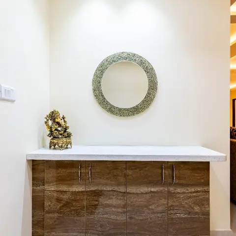Entryway console in wood-grain high-gloss finish with a white stone top, a round textured mirror above, and a brass Ganesha idol
