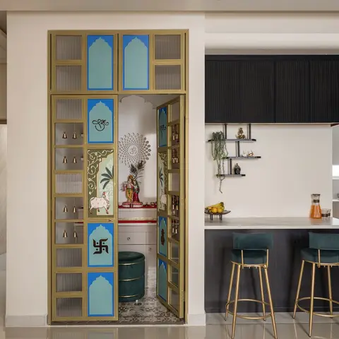 Entryway view combining a brass-framed glass pooja nook with a hand-painted Radha-Krishna panel alongside an open kitchen breakfast counter with teal velvet