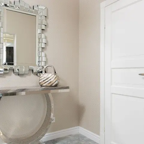 Entryway vignette with a faceted beveled-glass mirror above a round chrome console table, against dotted textured wallpaper beside a white panel door.