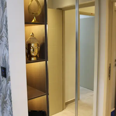 Entryway wardrobe wall with full-height mirror-panel doors beside an open backlit display niche styled with a gold Buddha head and decorative sculptures.