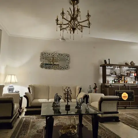 Evening shot of a traditional drawing room where a crystal-drop brass chandelier and a carved silver-leaf mirror hang over a cream teak sofa