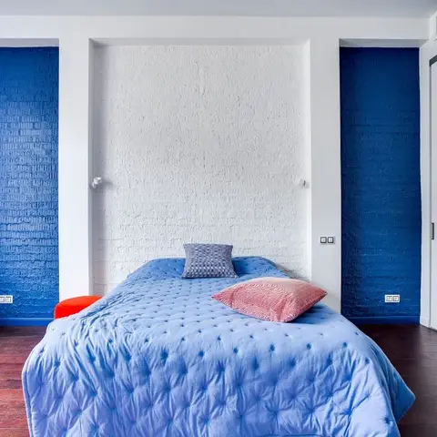 Exposed brick bedroom wall painted cobalt on both flanks with a white-painted brick alcove framing the bed, a blue tufted quilt and one coral cushion