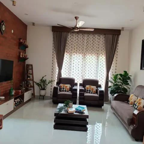 Exposed red-brick wall carrying the TV and a stepped display shelf of brass curios, facing two brown recliners and a leather sofa under block-print sheers.