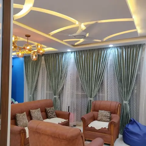 Family living room under a curved-cutout POP ceiling with concealed cove light, tan leatherette teak-frame sofas facing sage-green drapes over lace sheers