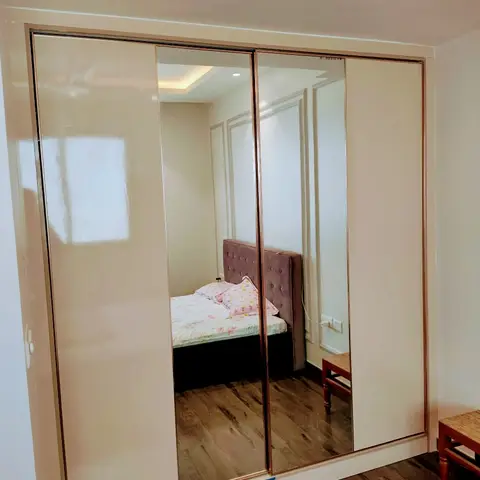 Floor-to-ceiling cream sliding wardrobe with rose-gold framed mirror shutters, the reflection showing the tufted grey bed and moulded wall behind the camera.