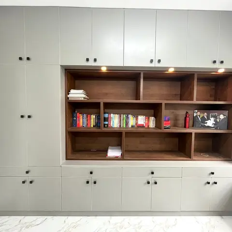 Floor-to-ceiling grey-shutter storage wall with an open wood-veneer shelving bay displaying books and framed photos, lit by small accent downlights.