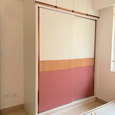 Floor-to-ceiling sliding wardrobe with cream, wood-grain, and dusty-pink laminate bands and a matching loft above, in a kids' bedroom.