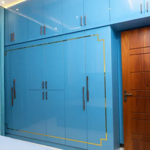 Floor-to-ceiling wardrobe in glossy cerulean lacquer outlined by a stepped brass inlay, with rose-gold bar handles and a grooved teak door alongside