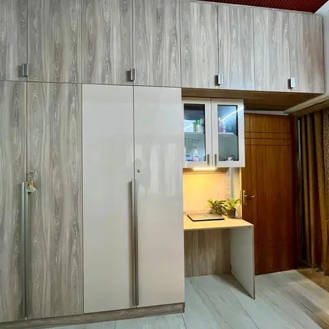 Floor-to-ceiling wardrobe wall in mixed grey woodgrain and glossy beige laminate, with an integrated study desk nook and glass-front overhead cabinet.