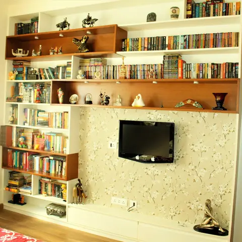 Floor-to-ceiling white bookshelf grid with teak-lined display boxes and shelf spotlights, packed with paperbacks and brass figurines