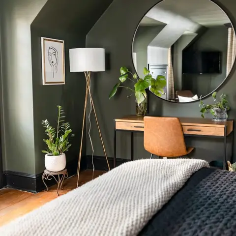 Foot of a bed in an olive-painted room with black skirting, a round black-framed mirror over a wood-and-steel desk and a tan leather swivel chair.
