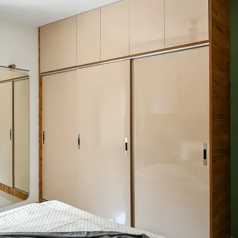 Foot of a quilted bed with a grey throw, facing a full-height beige high-gloss sliding wardrobe edged in teak with separate loft shutters
