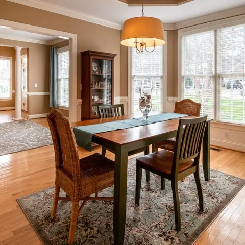 Formal dining room with a dark-green trestle table and mismatched wicker-and-wood chairs on a floral area rug, a drum-shade chandelier overhead