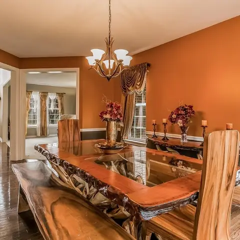 Formal dining room with a live-edge wood table with glass insert, burnt-orange walls, a candelabra chandelier, and draped swag curtains