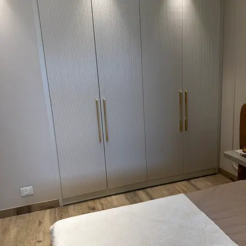Four-door fluted wardrobe in off-white recessed under a lit ceiling pocket with slim brass bar handles