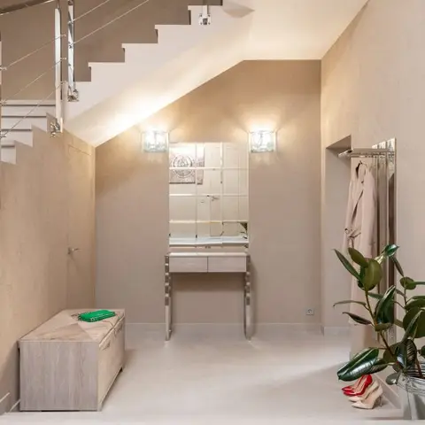 Foyer nook under a staircase with a mirrored console table, wall sconces, a wood storage bench, and a potted rubber plant by the entry.