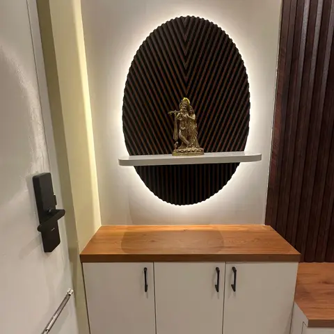 Foyer wall niche with a backlit oval fluted-wood panel framing a brass Radha-Krishna idol on a floating shelf, above a wood-topped white shoe cabinet.
