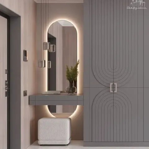 Foyer with a grey floor-to-ceiling cabinet routed in concentric arches, facing a backlit oval mirror over a slim grey drawer console and boucle cube stool.
