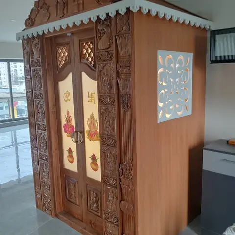 Free-standing carved wood-finish pooja mandir with backlit painted-glass doors depicting Ganesh and Om, set in the corner of an under-finished apartment.