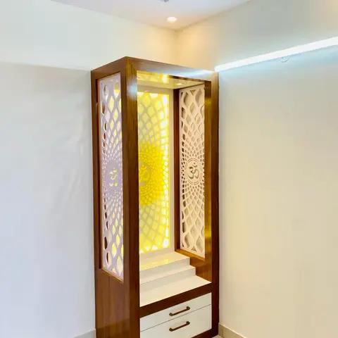 Free-standing corner pooja unit in wood veneer with carved jaali side panels bearing Om motifs, backlit yellow panel, marble steps, and a white drawer base.