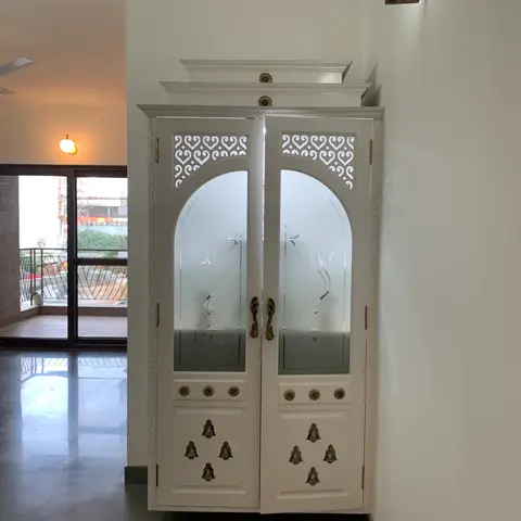 Free-standing white pooja unit with arched frosted-glass double doors, a carved jaali crown and brass bell-motif hardware