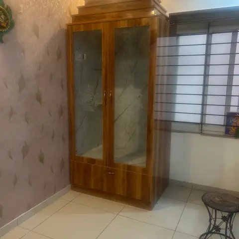 Free-standing wood-finish pooja cabinet with etched glass shutters and a temple-style pediment against wallpapered walls