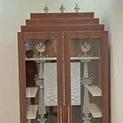 Free-standing wooden pooja cabinet with etched-glass doors showing a diya-and-peacock motif and a stepped pyramid top holding brass kalash finials.