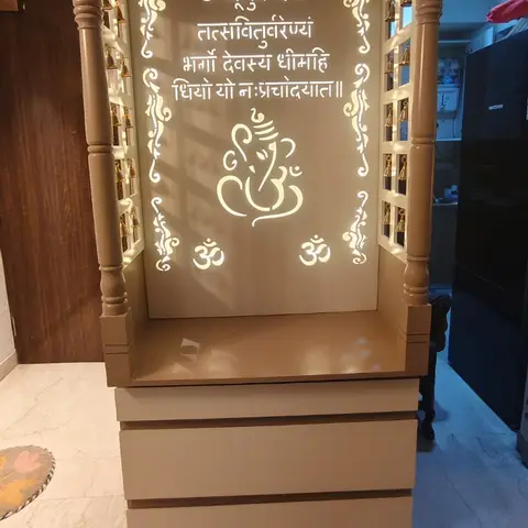 Freestanding beige pooja unit with a backlit Ganesha and Gayatri mantra panel, carved jali canopy and hanging brass bells, placed against a home entryway wall.